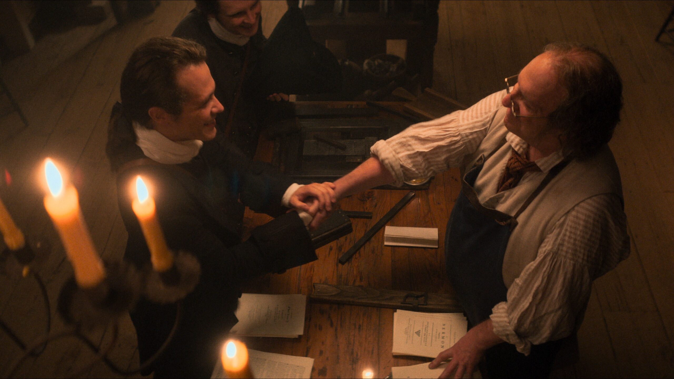 Jonathan Blair and John Paul Sneed portray Whitefield and Franklin clasping hands across a candlelit table with printed papers in A Great Awakening.